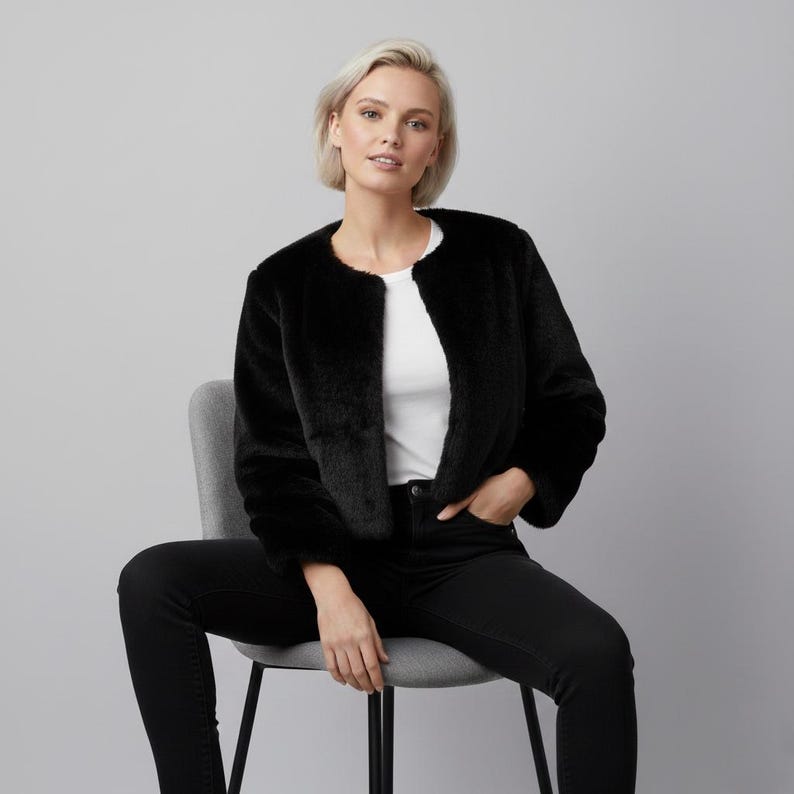 May include: A black faux fur jacket with a round neckline. The cropped jacket is worn over a white top and black jeans. The model is seated on a gray chair against a gray background.