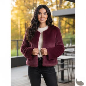 May include: A burgundy faux fur jacket with a round neckline and long sleeves. The jacket is open in the front, revealing a cream-colored sweater underneath. The person is wearing black jeans. The background is blurred with a natural outdoor setting.