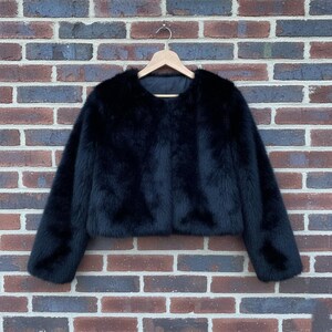 May include: A black faux fur jacket with a cropped silhouette hangs on a wooden hanger against a brick wall. The jacket has long sleeves and a round neckline, offering a luxurious and stylish look.