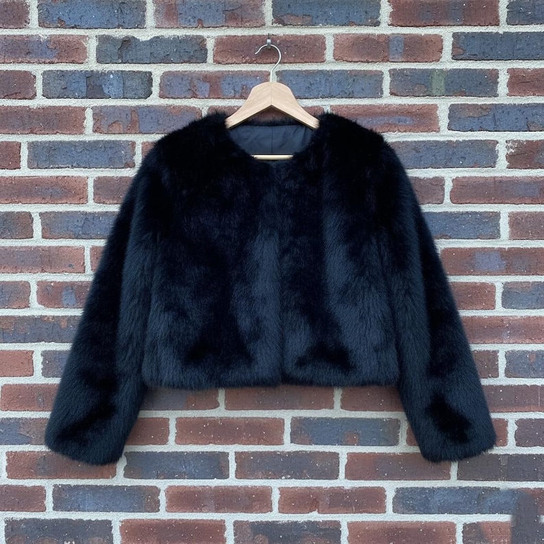 May include: A black faux fur jacket with a cropped silhouette hangs on a wooden hanger against a brick wall. The jacket has long sleeves and a round neckline, offering a luxurious and stylish look.