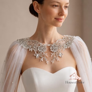 May include: A white bridal capelet featuring intricate lace detailing adorned with clear crystals, silver beads, and small white pearls. The sheer fabric drapes over the shoulders, and the piece includes the House of Befer logo text.