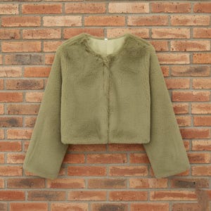 May include: A cropped, olive green faux fur jacket with long sleeves. The jacket has a round neckline and a simple, buttonless closure. The soft, textured fabric gives the jacket a plush appearance, suitable for cooler weather.