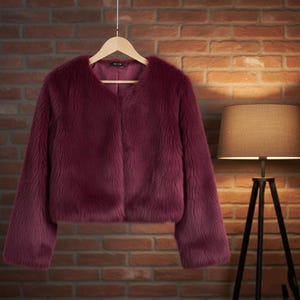 May include: A burgundy faux fur jacket with a V-neck and long sleeves hangs on a wooden hanger. The jacket is displayed against a brick wall, with a lamp in the background.