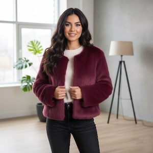 May include: A burgundy faux fur jacket with a round neckline and long sleeves. The jacket is open in the front and worn over a white sweater. The model is wearing black jeans. The background includes a window, a plant, and a floor lamp.