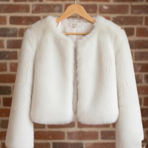 May include: A white faux fur jacket with a round neckline and long sleeves. The jacket is hanging on a wooden hanger against a brick wall background. The jacket has a soft, fluffy texture.