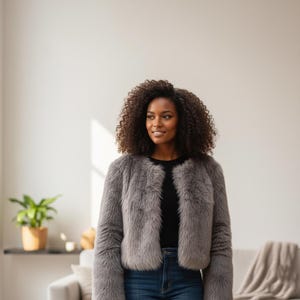 May include: A woman wearing a gray faux fur jacket over a black top and blue jeans. The jacket has a collarless design and a cropped length. The woman has curly dark hair and is smiling. The background is a neutral-toned living room.