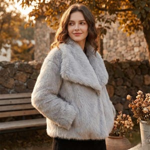 May include: A light gray faux fur coat with a wide collar. The coat has long sleeves and a soft, textured appearance. The woman in the image is wearing the coat outdoors, with a blurred background of trees and a stone wall.