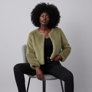 May include: A woman wearing a light olive green faux fur jacket with a round neckline and long sleeves. She is wearing a black top and black jeans. She is seated on a gray chair against a gray background.