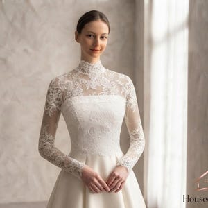 May include: A white floor-length gown featuring a high-neck lace bodice with long sheer lace sleeves and a solid satin skirt. The lace overlay displays intricate floral patterns extending from the neckline down to the fitted waistline.