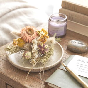 May include: A small, speckled ceramic dish holds a dried floral arrangement featuring a large pink strawflower, yellow billy balls, white statice, wheat stalks, and fluffy bunny tails. Nearby sits a lavender candle, a stone with the word COURAGE, and a pen.