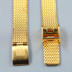 May include: A gold-tone metal watch strap featuring a fine mesh weave pattern. The strap includes a fold-over clasp mechanism with the Omega Greek letter symbol engraved on the surface. The metal has a polished finish and a consistent texture.