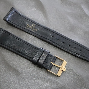 May include: A black leather watch strap featuring a gold-tone metal buckle with the Omega logo. The inner side of the longer strap segment displays the gold-printed text OMEGA Swiss Made. The strap has visible stitching along the edges.