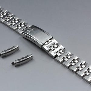 Puede incluir: Una correa de reloj plateada con un diseño de eslabones rectangulares. La correa incluye un cierre con el logotipo de Omega y dos piezas finales adicionales. La correa del reloj es de metal y se muestra sobre una superficie gris.