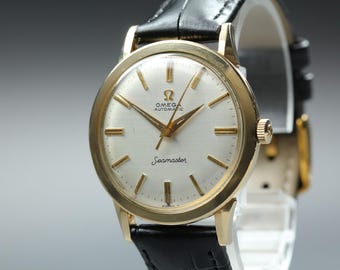 Può includere: Un orologio da polso Omega Seamaster Automatic con cassa rotonda color oro, quadrante bianco con indici e lancette color oro e cinturino in pelle nera. Il quadrante mostra il logo Omega, la parola Automatic e il nome Seamaster.