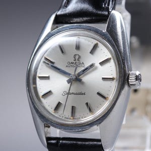 Könnte beinhalten: Eine silberne Omega Seamaster Automatik-Armbanduhr mit schwarzem Lederarmband. Das Zifferblatt der Uhr ist silberfarben mit schwarzen Zeigern und Markierungen. Die Uhr hat das Omega-Logo und "Seamaster" auf dem Zifferblatt. Die Uhr ist mit "Swiss Made" gekennzeichnet.