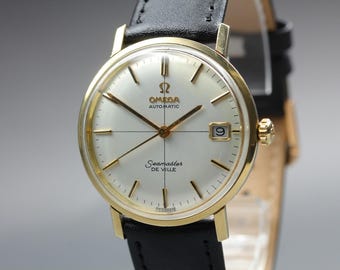 Può includere: Un orologio da polso automatico Omega Seamaster De Ville con cassa rotonda color oro, quadrante bianco con indici e lancette color oro, datario a ore tre e cinturino in pelle nera con fibbia color oro.