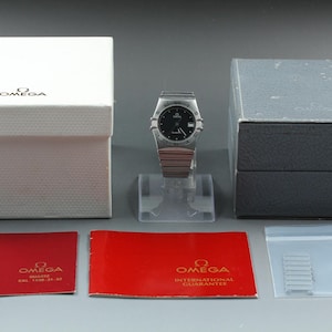 Könnte beinhalten: Eine Omega Constellation Armbanduhr mit schwarzem Zifferblatt und silberfarbenem Metallarmband, präsentiert mit einer weißen Außenbox, einer grauen Innenbox, zwei roten Omega-Anleitungsheften und einem kleinen Plastikbeutel mit Ersatzgliedern.