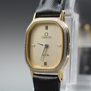 May include: An Omega De Ville wristwatch featuring a gold-toned textured dial with the Omega logo and De Ville text. The watch has a gold-toned bezel with a silver-toned textured border, silver-toned hands, and a black leather strap.