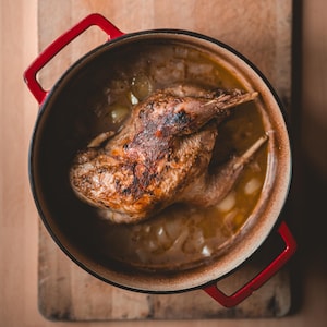May include: A roasted bird in a red cast iron pot with red handles, sitting on a wooden cutting board. The bird is golden brown, and the pot contains a light-colored broth with visible onions. The cutting board is light brown.