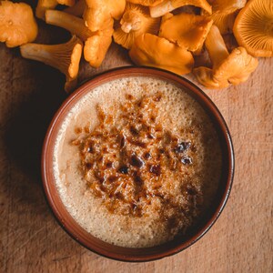 Könnte beinhalten: Draufsicht auf eine Schüssel mit cremefarbenem, hellbraunem Getränk mit karamellisierter Oberfläche, neben einer Ansammlung leuchtend orangefarbener Pfifferlinge. Die Anordnung befindet sich auf einem Holzbrett und wirkt rustikal und einladend.