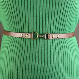 Pode incluir: Um cinto de couro castanho com fivela e rebites de latão. O cinto é usado sobre um vestido verde canelado. A palavra "PHAM" está estampada no cinto.