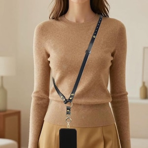 Puede incluir: Una correa cruzada para teléfono de cuero negro con herrajes dorados. La correa está sujeta a una funda de teléfono negra. La correa se lleva cruzada. La persona lleva un jersey y una falda color beige.