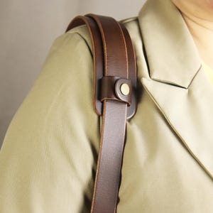 以下が含まれることがあります： 真鍮色のリベットが付いた、茶色の革製ショルダーストラップのクローズアップ。 ストラップはベージュのジャケットに取り付けられています。 革は高品質に見え、耐久性のあるデザインを示唆しています。