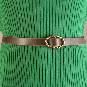 May include: A brown leather belt with a gold-colored oval buckle. The belt is worn over a green ribbed knit dress. The buckle has a twisted rope design.