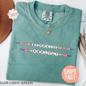 Op de afbeelding: Lichtgroen T-shirt met de woorden "BEHAVIOR ANALYST" in witte kralen, versierd met kleurrijke vlinder- en stermotieven. Een zilveren ketting met een hanger ligt op het shirt. Het shirt is gemarkeerd met "COLOR LIGHT GREEN".