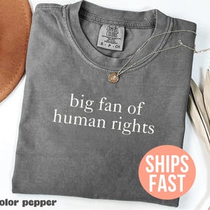Puede incluir: Una camiseta gris Comfort Colors con el texto blanco "big fan of human rights". Un collar con colgante dorado descansa sobre la camisa. La camisa está doblada y tiene cuello redondo.