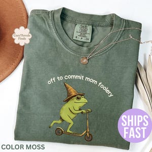 Puede incluir: Una camiseta verde musgo con la frase "off to commit mom foolery" y una rana de dibujos animados montada en un scooter. La rana lleva un sombrero de mago. También se ve un collar dorado y un sombrero marrón.