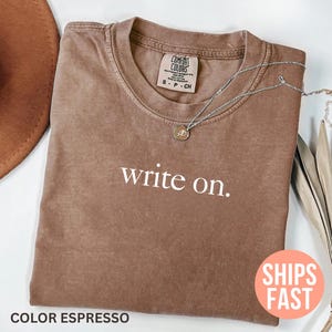 Puede incluir: Camiseta color espresso con la frase "write on" en blanco. La camiseta tiene cuello redondo y mangas cortas. Un collar plateado con un pequeño colgante descansa sobre la camiseta. La camiseta es parte de una colección Comfort Colors.