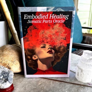May include: A book titled "Embodied Healing Somatic Parts Oracle" features a woman with red lips and a red cloud-like design above her head. The book is on a table with crystals and other objects.
