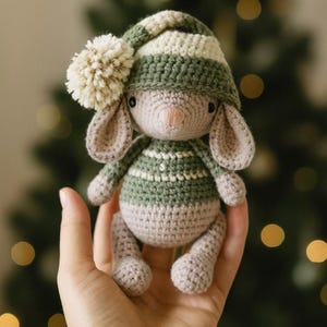 May include: A handmade crochet bunny doll with a green and white striped hat and sweater. The bunny has long ears, a pink nose, and a cream-colored pom-pom on its hat. The doll is made of soft yarn and is a cute, cuddly toy.