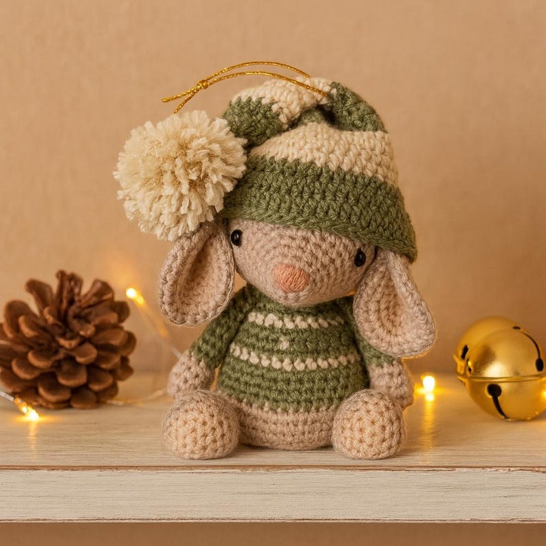 May include: A handmade crochet bunny doll wearing a green and white striped hat and sweater. The bunny has long ears, a beige pom-pom, and a pink nose. The doll is sitting on a white surface with a pinecone and gold bells in the background.