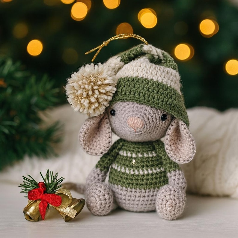 May include: A crocheted mouse wearing a green and white striped hat and sweater, with a gold pom-pom. The mouse has large ears and is sitting next to gold bells with a red bow and greenery. The background has blurred Christmas lights.