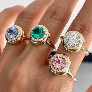 Puede incluir: Cuatro anillos de oro, cada uno con una gran piedra central de corte redondo en azul claro, verde esmeralda, blanco transparente y rosa suave. Cada piedra está rodeada por un halo de pequeños diamantes pavé en una banda de oro pulido.
