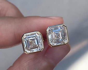 May include: A pair of square-cut diamond stud earrings with a gold bezel setting. The diamonds have a brilliant, multifaceted appearance, reflecting light. The earrings are a classic jewelry item.