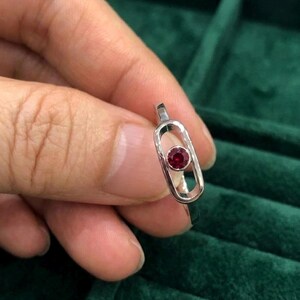 May include: A silver ring featuring a small, round, red gemstone set within an elongated, open oval design. The ring is held against a dark green velvet background, highlighting the jewelry's details.