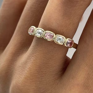 Pode incluir: Um anel de banda dourada com cinco pedras preciosas de corte almofada engastadas em bisel e dispostas em fila. As pedras alternam entre três gemas cor-de-rosa e duas gemas brancas transparentes, fixadas em monturas douradas lisas e arredondadas.
