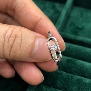 May include: A silver ring featuring a small, round diamond set within an elongated, open oval design. The ring is held against a dark green velvet surface, showcasing its delicate details and elegant simplicity.