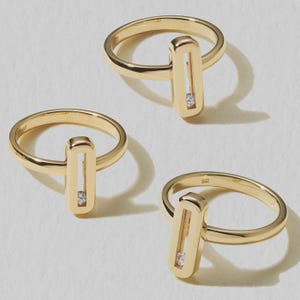 Op de afbeelding: Drie gouden ringen met een rechthoekig ontwerp met een open midden en een kleine, heldere steen. De ringen worden gepresenteerd op een wit oppervlak, waarbij hun gepolijste afwerking wordt benadrukt. Minimalistisch sieradenontwerp.