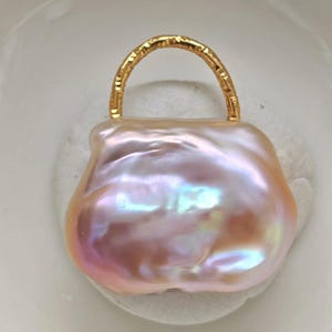 May include: A lustrous, baroque pearl pendant in the shape of a handbag. The pearl has a soft pink hue with iridescent flashes of color. A textured gold-colored handle arches over the top, completing the design.