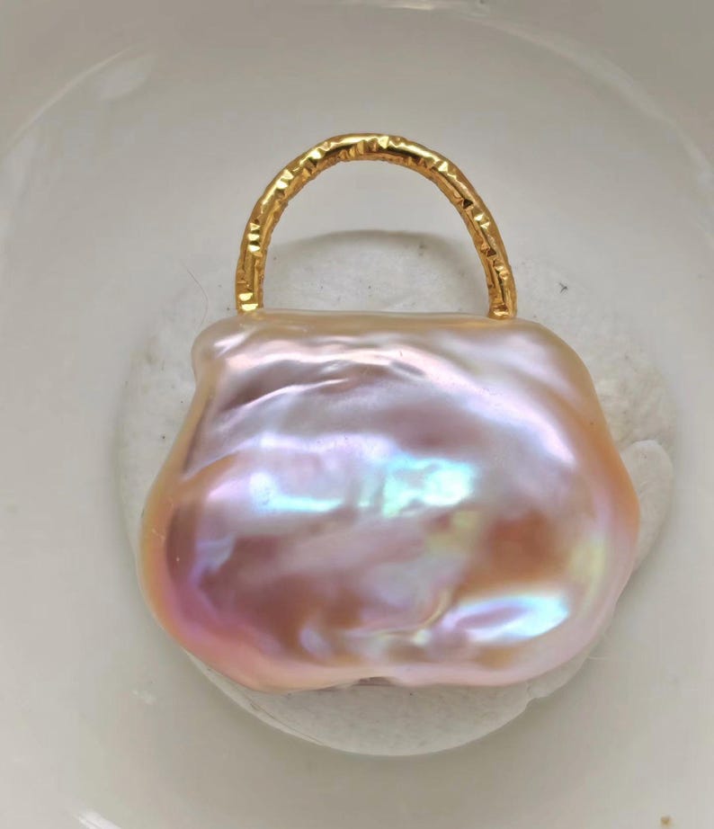 May include: A lustrous, baroque pearl pendant in the shape of a handbag. The pearl has a soft pink hue with iridescent flashes of color. A textured gold-colored handle arches over the top, completing the design.