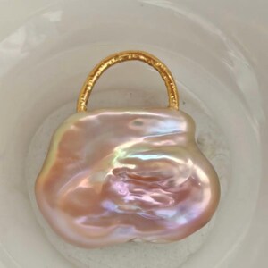 May include: A large, irregularly shaped, iridescent pink pearl pendant with a gold-colored handle. The pearl has a smooth, polished surface with a subtle sheen, and the handle is a curved, textured metal. The pendant is set against a white background.