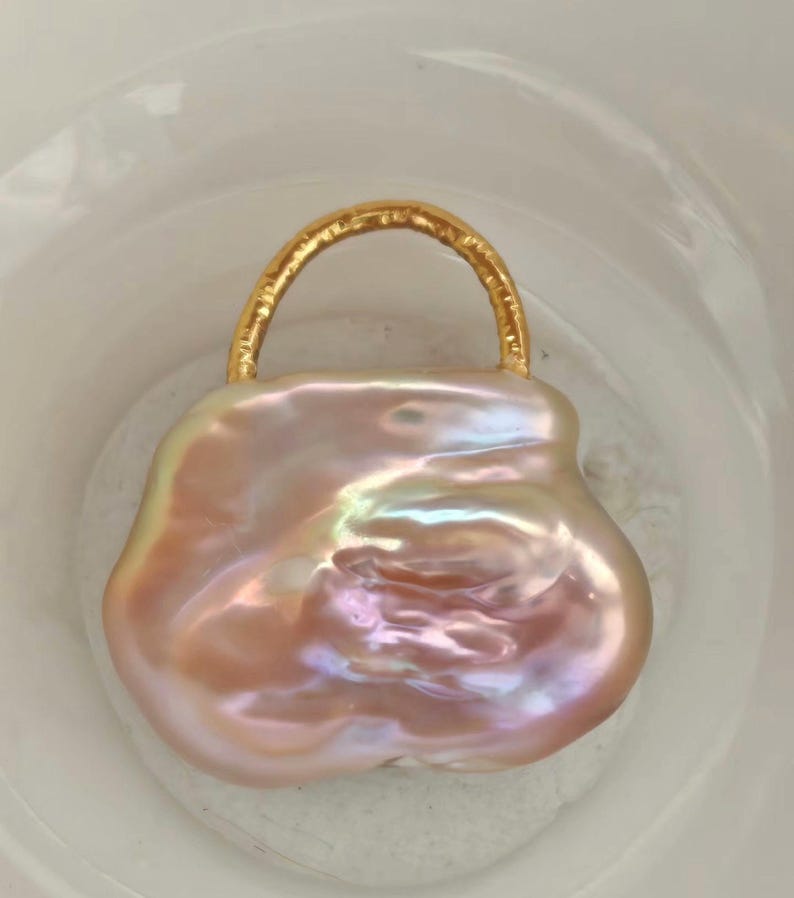 May include: A large, irregularly shaped, iridescent pink pearl pendant with a gold-colored handle. The pearl has a smooth, polished surface with a subtle sheen, and the handle is a curved, textured metal. The pendant is set against a white background.
