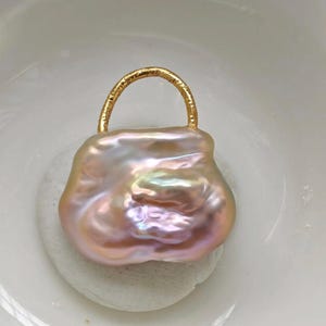 May include: A unique, baroque pearl pendant in the shape of a handbag. The iridescent pearl has pink, peach, and lavender hues. A gold-colored metal handle arches over the top of the pearl, creating a stylish jewelry piece.