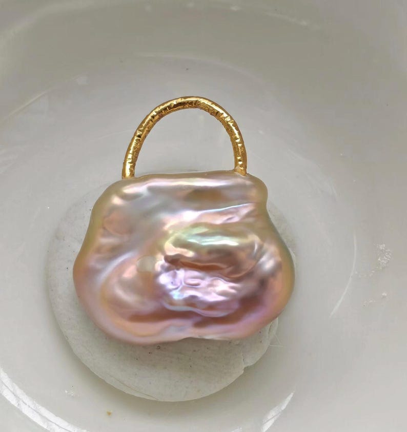 May include: A unique, baroque pearl pendant in the shape of a handbag. The iridescent pearl has pink, peach, and lavender hues. A gold-colored metal handle arches over the top of the pearl, creating a stylish jewelry piece.
