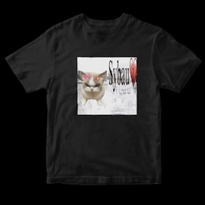 Op de afbeelding: Zwart T-shirt met een afbeelding van een kat met een roze strik en een rode ster. Het woord "Sybau" is gedrukt in een gestileerd lettertype, met een gebroken hart. Het T-shirt is gemaakt van zacht materiaal.