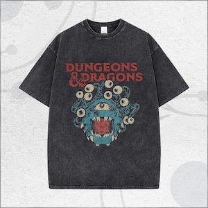 May include: Dark gray t-shirt featuring the text "DUNGEONS & DRAGONS" in red. The shirt has a distressed look and a graphic of a blue monster with multiple eyes and a red die in its mouth.